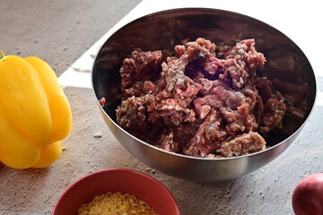 Raw ground beef in a silver bowl surrounded by fresh vegetables on a light background. A clean, bright setup representing home cooking, meal preparation, and fresh ingredients