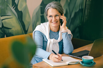 Confident older woman talking on smartphone while working with laptop and notebook in cafe