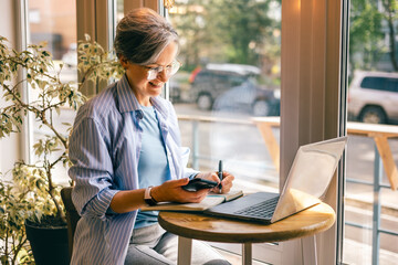 Senior businesswoman writing notes from phone at cafe table with laptop, smiling, working remotely