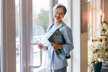 Confident senior woman holding laptop and entering office building with smile and natural light