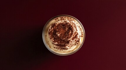 An overhead shot of classic tiramisu dusted with cocoa powder in a glass cup placed on a solid deep burgundy background creamy textures 
