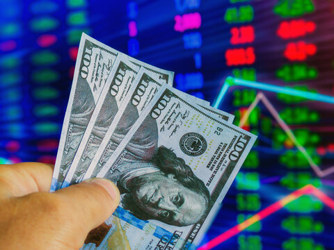 A hand holding several hundred-dollar bills is positioned in front of a colorful stock market display showing fluctuating values and trends.