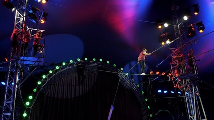 A brave tightrope walker walks on a tightrope during a circus show, he holds a long stick in his hands for balance and dances. Tightrope walkers performing under the light of multicolored spotlights.
