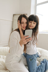 A mother and daughter are sitting together, looking at a phone while having a video call. They smile warmly, clearly enjoying the moment and the person on the other end of the screen. 