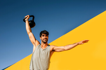 Hispanic athlete performs kettlebell exercise against bright yellow backdrop during outdoor workout...