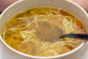 Clear chicken broth with thin noodles, carrot slices, and parsley served in a bowl. A spoon rests inside the hot, comforting soup.
