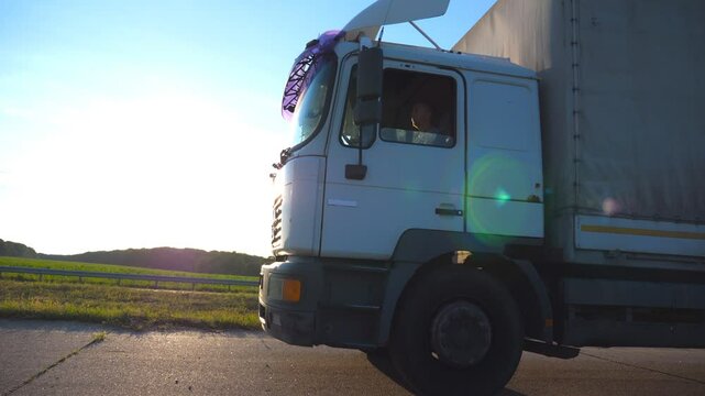 Camera follow to truck driving on a highway. Lorry rides through the countryside with sun shine at background.Wheels of truck ride at asphalt. Driver looks carefully at the road. Side view