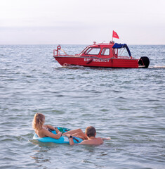 A young couple swimming in the sea with an inflatable circle watches as a red rescue boat passes by them