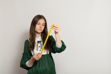 A woman wearing a green dress inspects a yellow measuring ruler while standing indoors against a...