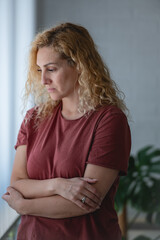 Thoughtful woman standing by a window with arms crossed, looking down with a serious expression, reflecting or feeling sad in a quiet home environment.