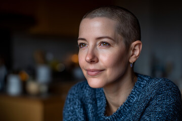 Woman with shaved head wearing blue sweater, looking to the side in warm candle-lit room, symbolizing cancer awareness, resilience, inner strength, healing, and emotional peace in cozy setting