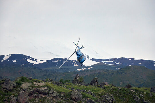 blue helicopter is maneuvering through the air above rocky terrain and lush green hills, with majestic snow-capped mountain peaks visible in the distance on a cloudy day