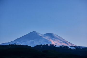 Snow-covered mountain peaks rise majestically against a clear blue sky as dawn approaches, casting soft light on the pristine snow. This serene landscape showcases natures beauty
