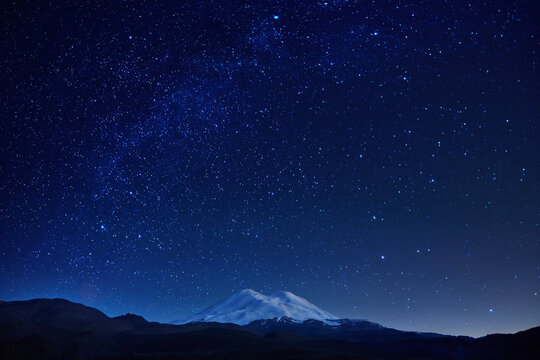 breathtaking view of a mountain with a snow-covered summit set against a clear night sky filled with vibrant stars and a rich backdrop of deep blue hues, creating a serene atmosphere