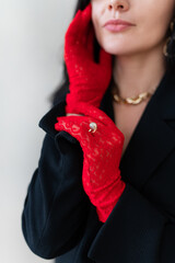 Elegant woman in red lace gloves and black blazer poses, touching face. Close-up highlights gold diamond ring over glove, gold necklace. Fashion, style, luxury accessories