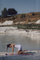 Woman in white sportswear lifting leg in kneeling position on yoga mat by lake. Outdoor yoga and body conditioning concept. Design for poster, banner, greeting card, invitation. Side view