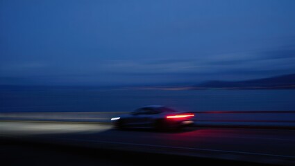 Blurred modern car driving along a coastal road at dusk with headlights and taillights glowing against a dark blue sky and calm sea