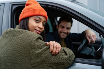 Woman wearing an orange beanie and olive jacket leaning on a car window smiling confidently with a man sitting in the driver seat smiling behind her