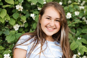 Smiling teenage girl 13 -14 year old with long healthy blonde hair over blooming jasmine bush with white flowers in garden outdoors. Happiness.