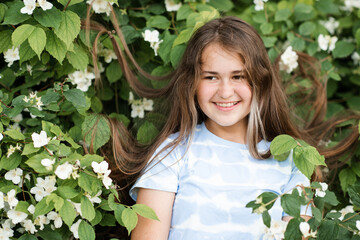 Smiling teenage girl 13 -14 year old with long healthy blonde hair over blooming jasmine bush with white flowers in garden outdoors. Happiness.