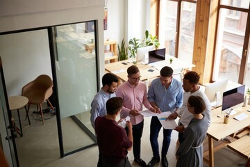 Group of six colleagues standing in a modern office discussing charts and documents with engaged and collaborative expressions