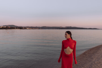 Woman in red cut-out dress standing on beach at sunset. Fashion photography. Summer vacation and travel concept. Design for poster, banner, greeting card, invitation. Side view with copy space