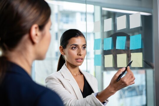 Two professional women collaborating and discussing ideas using colorful sticky notes on a glass wall in a modern office