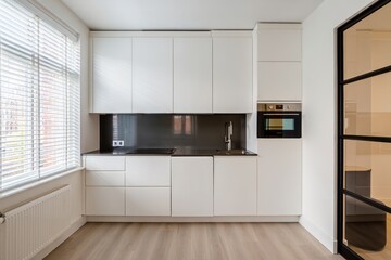 Fototapeta premium bright modern kitchen interior with white cabinets, black backsplash, built-in oven, large window with blinds, and wood flooring creating a clean and minimalist atmosphere