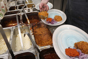 Chef serving rice and vegetable stew onto plates with red pepper relish and sliced onions in a restaurant kitchen. Food prep in progress at a buffet-style serving station.
