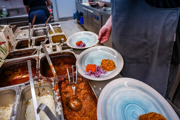 Chef preparing dishes in a restaurant kitchen with stews, sauces, rice, and fresh garnishes. Plates are being assembled with red sauce, sautéed vegetables, and sliced onions.