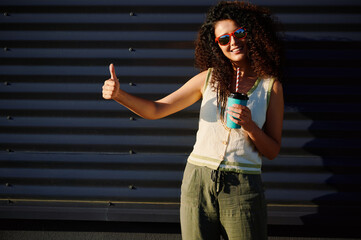 woman with sunglasses holds cup and shows thumbs-up gesture outdoors. casual summer style, joyful...
