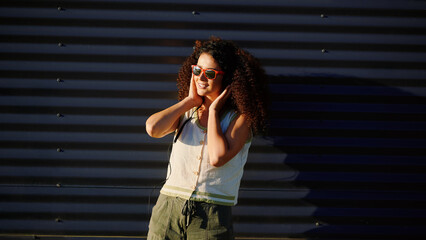woman enjoying sun wearing sunglasses against striped shadowed wall. curly-haired model in casual...