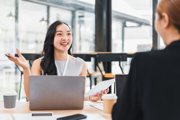 Two young Asian women having friendly conversation in modern cafe, one holding coffee cup and other using digital tablet, natural light and casual business attire