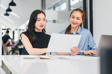 Obraz premium Two Asian women working together in modern office, discussing documents with laptop on table, showing teamwork and collaboration in bright workspace