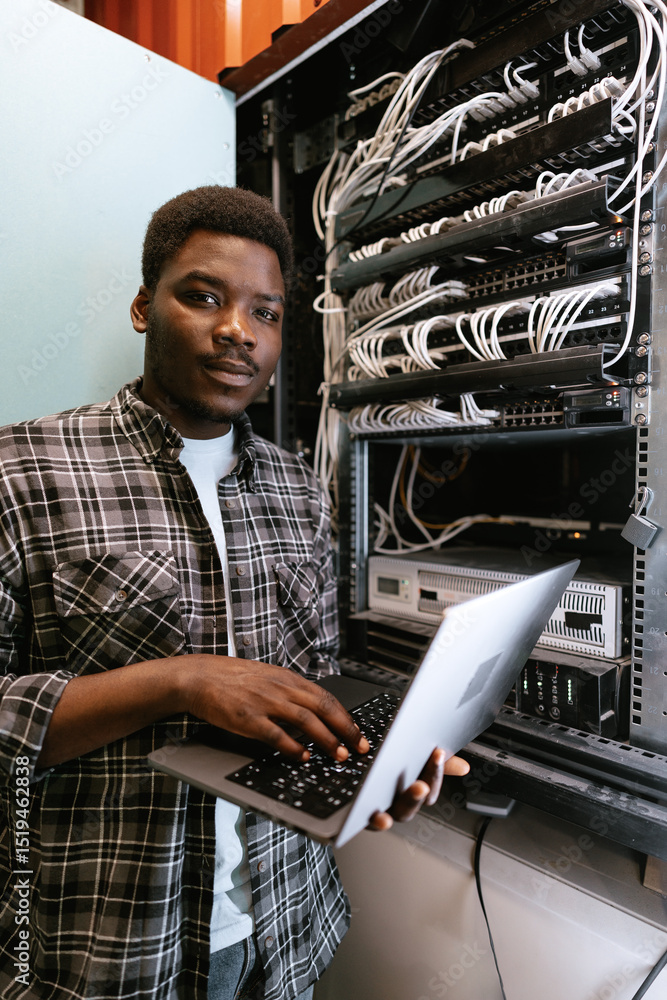 Poster man working on a laptop in a server room during daylight hours, managing network equipment and data 