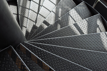 Curved metal staircase with textured diamond plate steps spirals downward, illuminated by soft daylight and dynamic shadows in a modern industrial interior.