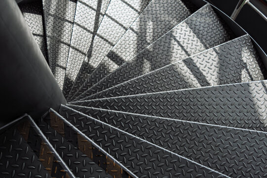 Curved metal staircase with textured diamond plate steps spirals downward, illuminated by soft daylight and dynamic shadows in a modern industrial interior.