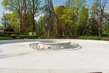 Circular dry fountain with central platform and metal fixtures sits in a landscaped city park, surrounded by green trees and wooden benches on a sunny day.