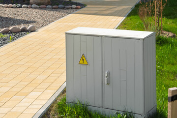 Metal electrical cabinet with warning sign stands on fresh green grass near modern paved walkway in urban landscaped area.