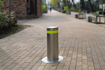 Stainless steel security bollard with yellow reflective stripe stands on paved pedestrian walkway in city park to control vehicle access.