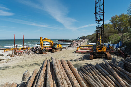 Yellow construction equipment and timber piles at a coastal site, with machinery working on shoreline reinforcement under a clear blue sky.