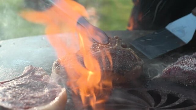 Close-up of juicy ribeye steaks being flipped over open flame on hot grill plate &ndash; barbecue cooking scene