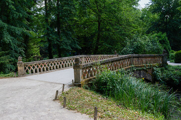 A rustic stone bridge with intricate railings spans a small stream in a lush, green park setting.