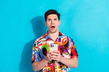 Young man in a vibrant colorful shirt holding smartphone and expressing amazement against a solid...