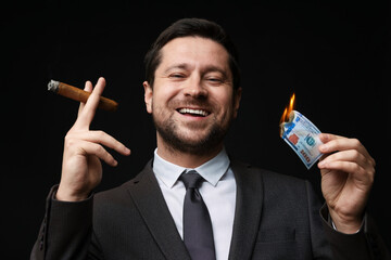 Happy greedy banker with burning dollar banknote smoking cigar on black background