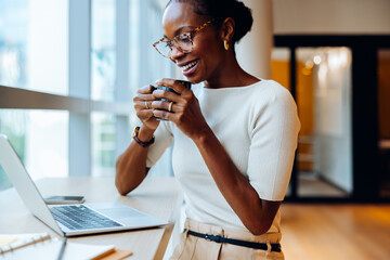 Woman enjoying coffee while working on a laptop in bright office setting
