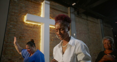 Woman clasping hands in prayer inside a church, expressing gratitude and faith, standing in front...