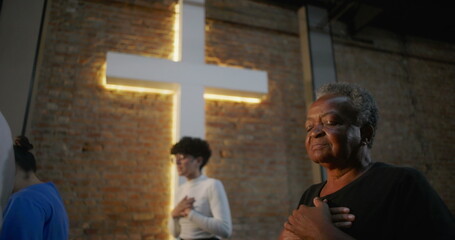 Elderly woman in church placing hands on chest, eyes closed in prayer, illuminated cross and brick...