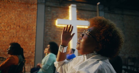 African American woman raising hand in prayer during worship service, eyes closed in deep spiritual...