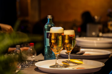 Two glasses of cold draft beer with foam served on a white tray in a cozy restaurant or bar. Warm ambient lighting and blurred background create a relaxed evening atmosphere.